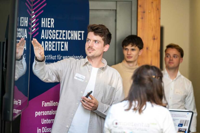 Fabian Düesmann Larocque (Head of People & Culture) is standing in front of a roll-up banner bearing the words “EXCELLENT WORK HERE” and is speaking to a small group. He is holding a dark pen in one hand and gesturing with the other.