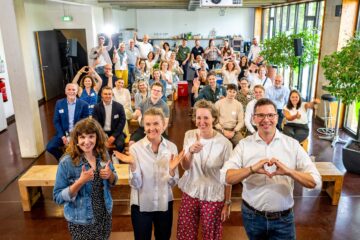 A large group photo in a bright function room: around 40 people are arranged in several rows – some standing at the front, others sitting on wooden benches behind them, and others standing at the very back. Four people in the foreground are particularly prominent; numerous are forming heart shapes with both hands, whilst others are giving a thumbs-up.
