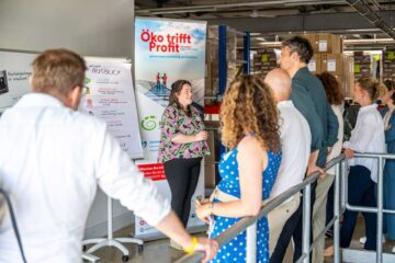 Ms Henriette Grunwald, wearing a colourful patterned top, is standing in front of a roll-up banner bearing the slogan “Eco meets profit – working together for sustainability” and is speaking to a group of at least seven people standing facing her. The room has a warehouse-like feel, with exposed shelving in the background.
