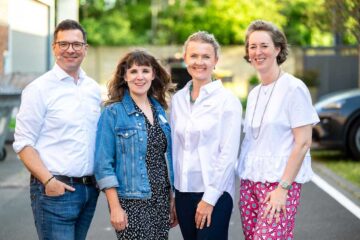 Four speakers are standing side by side in an outdoor car park, looking into the camera: a man (Mark Sethe) on the left, wearing a white shirt and jeans; next to him, a woman wearing a denim jacket over a patterned dress; then a woman in a white blouse with turquoise jewellery; and on the right, a woman in a white top and a patterned skirt.