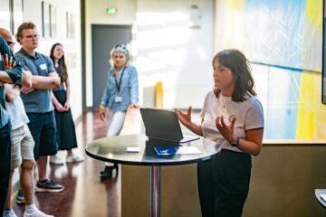 A woman with shoulder-length brown hair is speaking at a round bar table on which a tablet is open. She is gesturing with both hands. To her left are at least four people standing with their backs to the camera. In the background is a brightly lit corridor featuring a large blue and yellow mural.