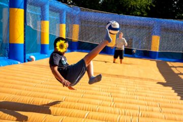 A child wearing a black T-shirt and dark shorts is performing a bicycle kick on an inflatable yellow-and-blue court, hitting a blue-and-white volleyball. In the background, a second person is standing behind a net.