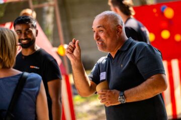 A middle-aged man in a dark polo shirt with a name badge is gesturing as he speaks; to his left is a smiling younger man with a beard; on the far left is a woman with a shoulder bag.