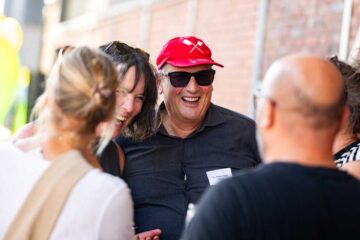 In the centre of the picture, Dr Klaus Schulz is smiling, wearing a red cap and dark sunglasses; on his black shirt is a name badge bearing the inscription ‘Dr Klaus Vitalis’. To his left, a woman with dark hair is smiling; other people are visible, some partially obscured, in the foreground and background.