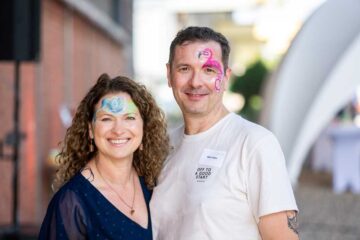 Mark Sethe and his wife are posing with smiles on their faces in an outdoor setting: a woman with curly hair and a blue top, wearing blue floral face paint; a man in a white T-shirt with pink flamingo face paint