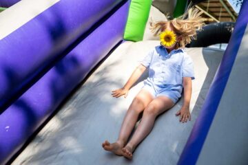 A child with blonde hair slides down an inflatable slide; a yellow sunflower covers their face, and their hair is blown upwards by the movement.