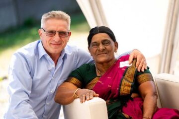 Two people are posing side by side for a photograph: an elderly man with grey hair, glasses and a light blue shirt has his arm around an elderly woman wearing a green and pink sari with gold trim and a gold bangle.