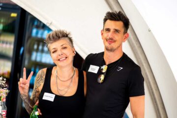 Two people wearing name badges in front of a drinks fridge: on the left, a woman (Evangelista) with short grey-blonde hair, tattoos and a black top, making a victory sign; on the right, a man with dark hair (Jonny), wearing a black Gymshark T-shirt and sunglasses on a chain around his neck.