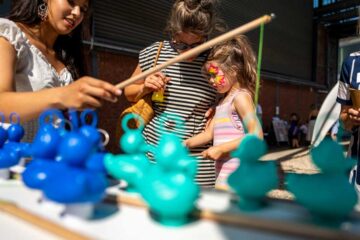 Fishing game: A child with colourful face paint and a woman in a striped dress are leaning over a table covered with blue and turquoise rubber figures; to the left is another woman in a white dress, pointing at the figures with a wooden stick.