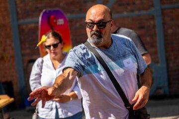 A bald man wearing sunglasses, a name badge and carrying a shoulder bag is throwing a dart at a target.