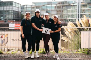 Team Glow & Row at the ‘Düsseldorf am Ruder’ charity regatta: Julia Kötting, Fred Kötting, Lynn-Marie Zapp and Pia Mertens are standing side by side in front of the banner of the Germania Düsseldorf Rowing Club; Lynn-Marie Zapp is holding start number 8. They are all wearing dark polo shirts and white caps.