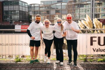 Piotr Jankiewicz, Marta Dorozinski, Daria Kruk and Dimitrij Michel pose in white ‘Schulz & Sohn’ T-shirts and colourful party glasses in front of a fence bearing the inscription ‘RCLUB GERMANIA DÜSSELDORF’.