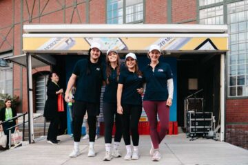 Team Glow & Row at the ‘Düsseldorf am Ruder’ charity regatta: four people in dark blue polo shirts and white caps are standing in front of the organiser’s stage; in the background, a brick façade and equipment cases.