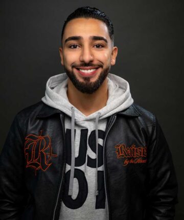 Ahmed hat dunkle Haare und trägt einen kurzen Bart, er lächelt sympathisch in die Kamera. Er trägt einen grauen Hoodie und darüber eine schwarze Lederjacke.