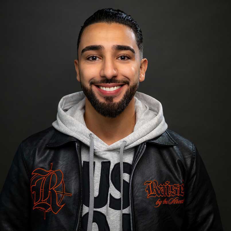 Ahmed hat dunkle Haare und trägt einen kurzen Bart, er lächelt sympathisch in die Kamera. Er trägt einen grauen Hoodie und darüber eine schwarze Lederjacke.