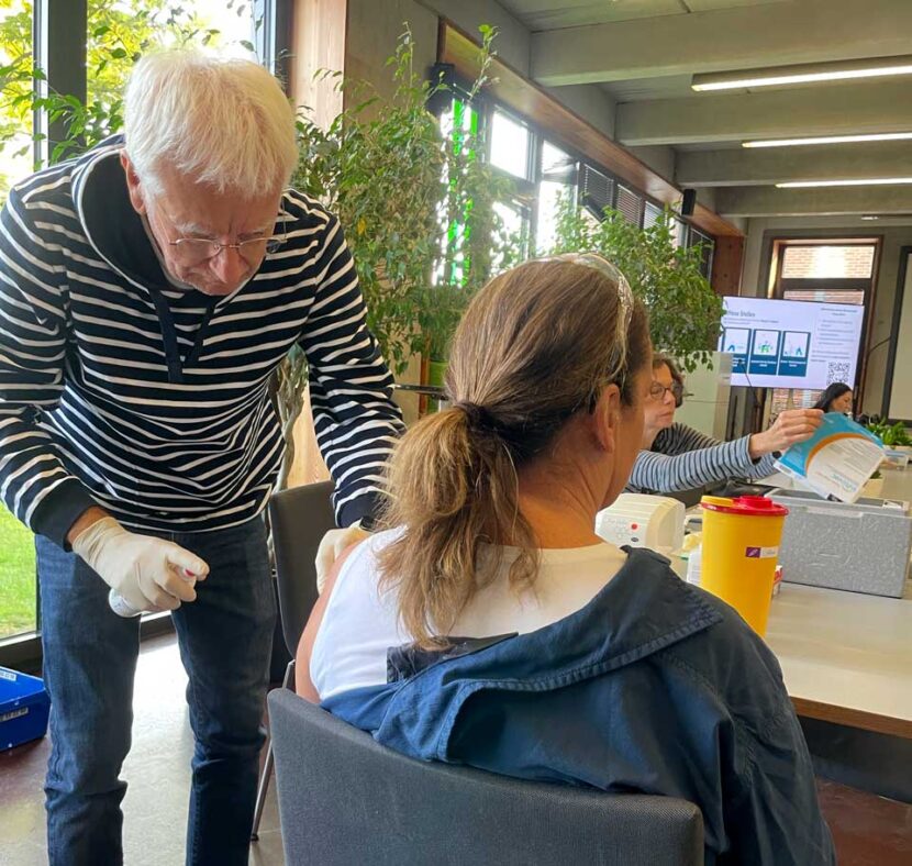Ein älterer Mann mit medizinischen Handschuhen sprüht aus einer kleinen Flasche auf den freigelegten Oberarm einer sitzenden Frau. Im Hintergrund sitzen zwei weitere Personen an einem langen Tisch mit einem gelben Behälter.